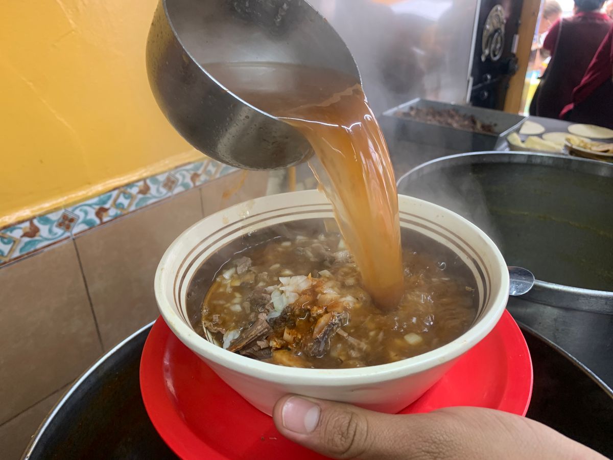 Cucharón sirviendo consomé caliente sobre un tazón de birria, con vapor, en el mostrador de la cocina