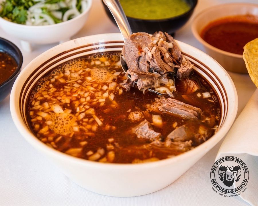 Plato de birria de chivo en su caldo, con cebolla picada y una cuchara con carne encima