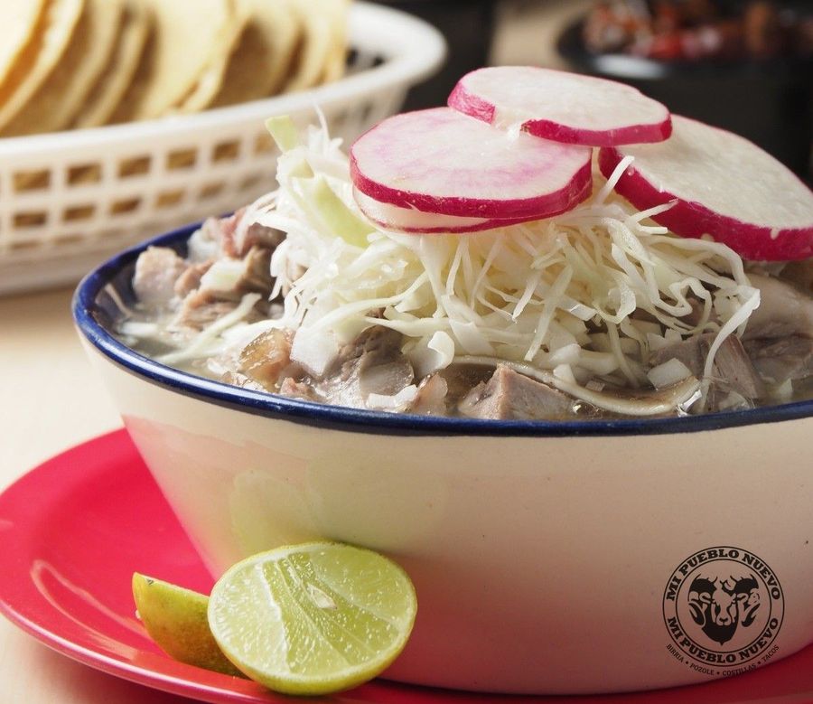 Tazón de pozole blanco con col, rábanos y limón, sobre plato rojo, con canasta de tortillas al fondo